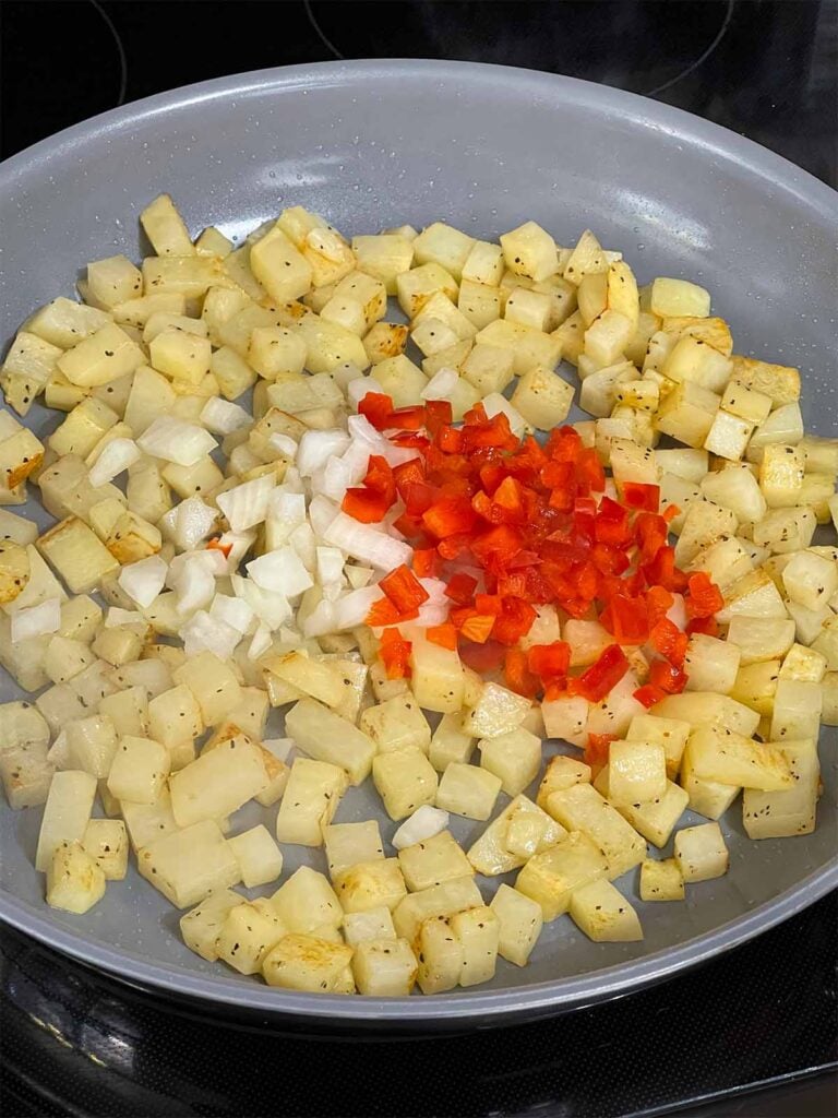 Onions and red bell pepper added to seasoned diced potatoes in a skillet.