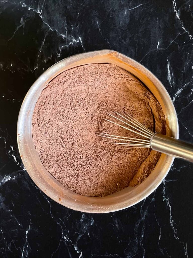 Dry ingredients for chocolate bundt cake whisked together in a metal mixing bowl on a dark surface.
