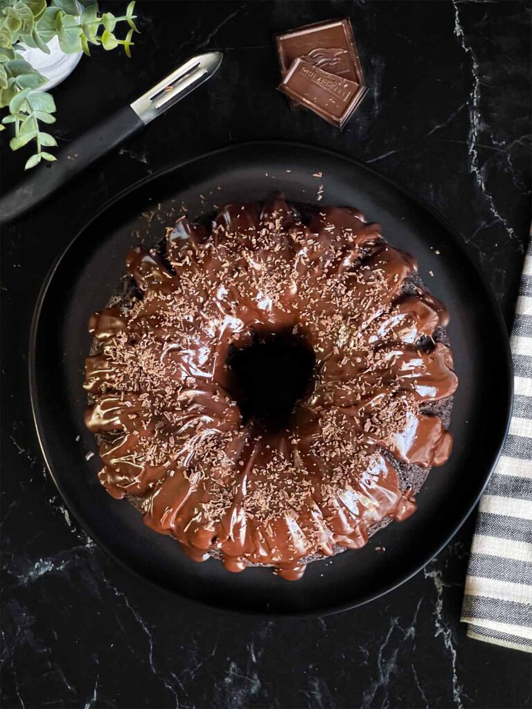 Chocolate bundt cake topped with chocolate ganache and garnished with chocolate shavings on a dark plate on a dark surface.