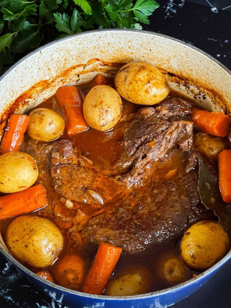 Classic pot roast with potatoes and carrots in a dutch oven on a dark surface.