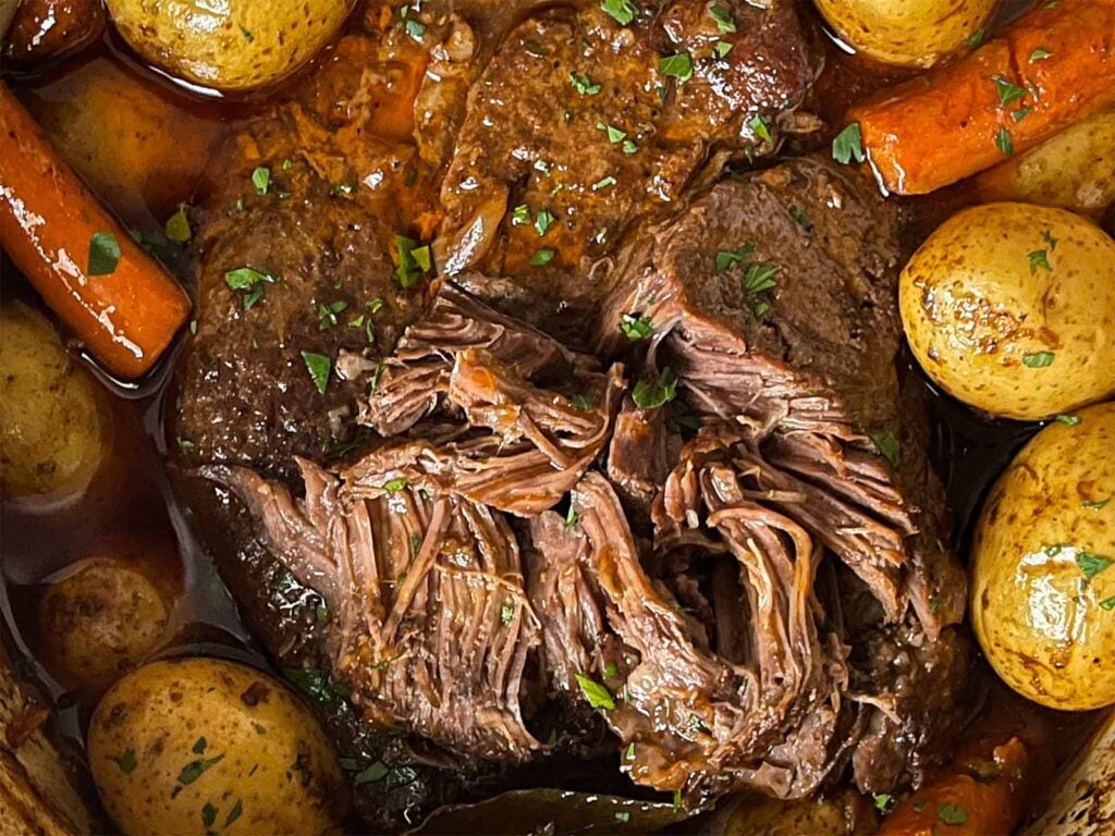 Classic pot roast with potatoes and carrots in a dutch oven on a dark surface.