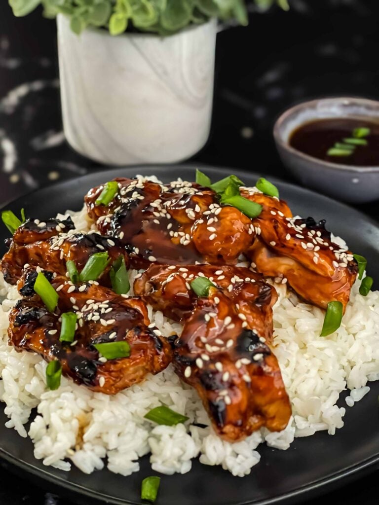 Bourbon chicken on a bed of white rice on a dark plate, garnished with sesame seeds and green onion.