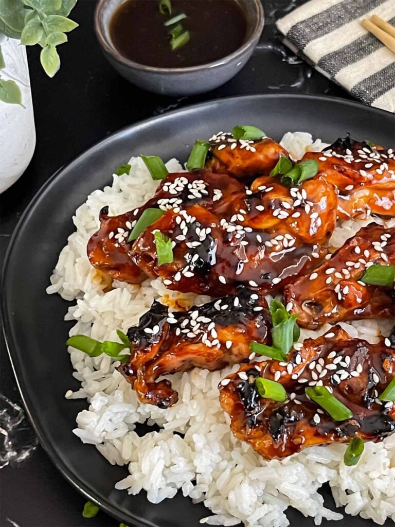 Bourbon chicken on a bed of white rice on a dark plate, garnished with sesame seeds and green onion.