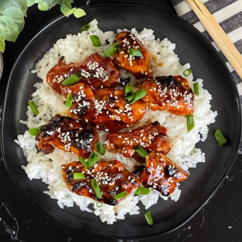 Bourbon chicken on a bed of white rice on a dark plate, garnished with sesame seeds and green onion.