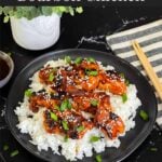 Bourbon chicken on a bed of white rice on a dark plate, garnished with sesame seeds and green onion.