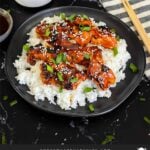 Bourbon chicken on a bed of white rice on a dark plate, garnished with sesame seeds and green onion.