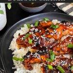 Bourbon chicken on a bed of white rice on a dark plate, garnished with sesame seeds and green onion.