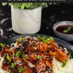 Bourbon chicken on a bed of white rice on a dark plate, garnished with sesame seeds and green onion.