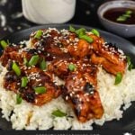Bourbon chicken on a bed of white rice on a dark plate, garnished with sesame seeds and green onion.