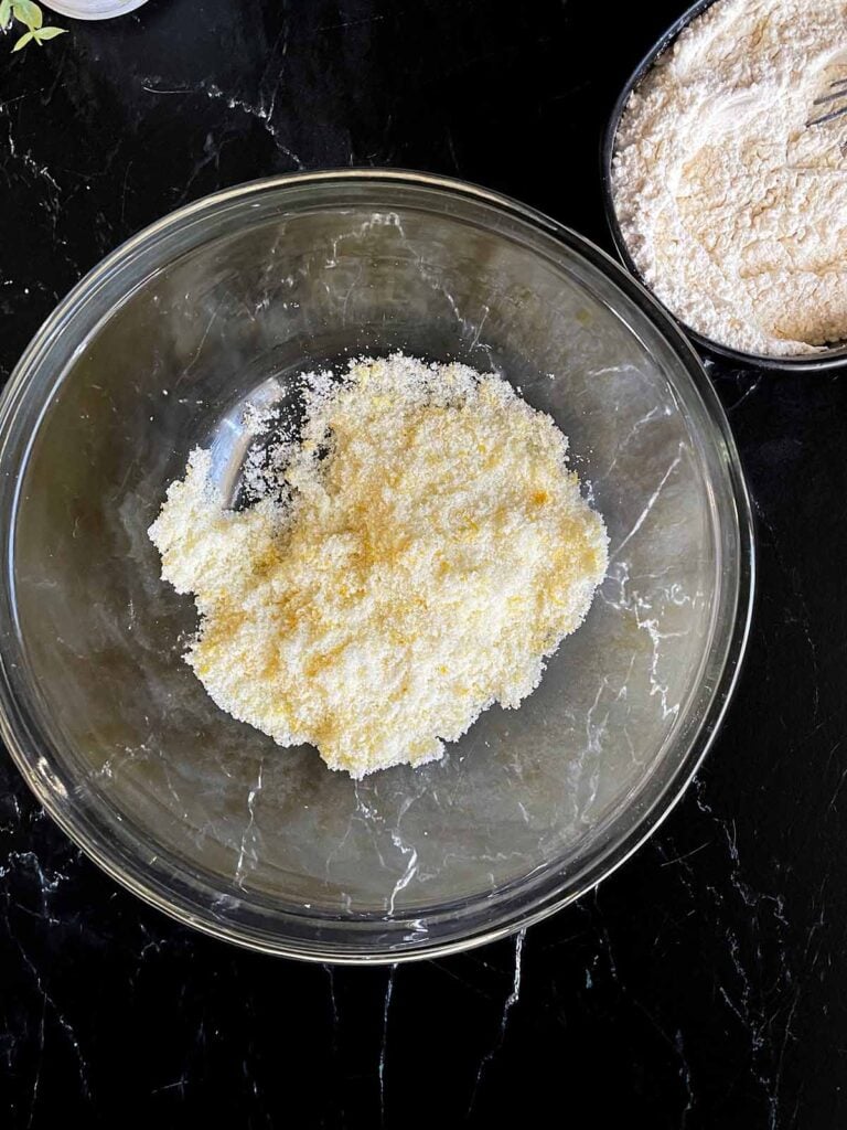 Granulated sugar and lemon zest rubbed together in a glass mixing bowl on a dark surface.