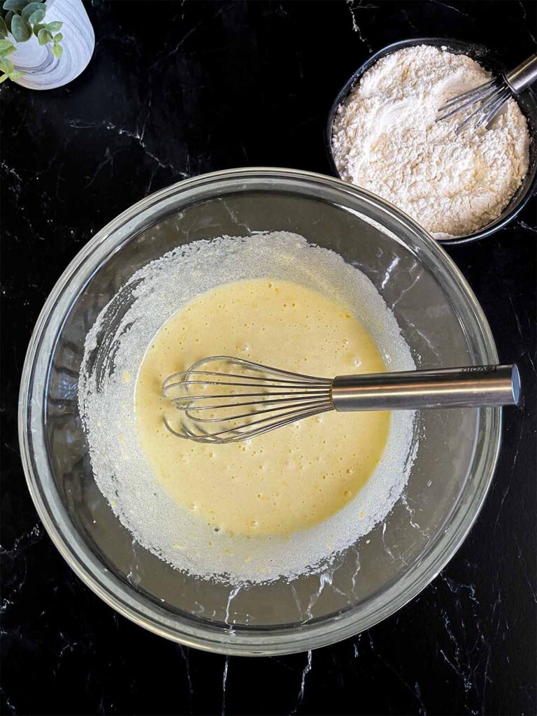 Eggs whisked into the lemon zest infused granulated sugar in a glass bowl on a dark surface.