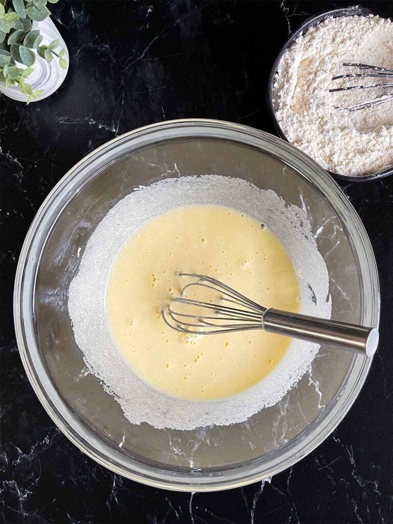 Eggs and granulated sugar beating until pale and thick in a glass mixing bowl with the dry ingredients in a dark bowl on a dark surface.