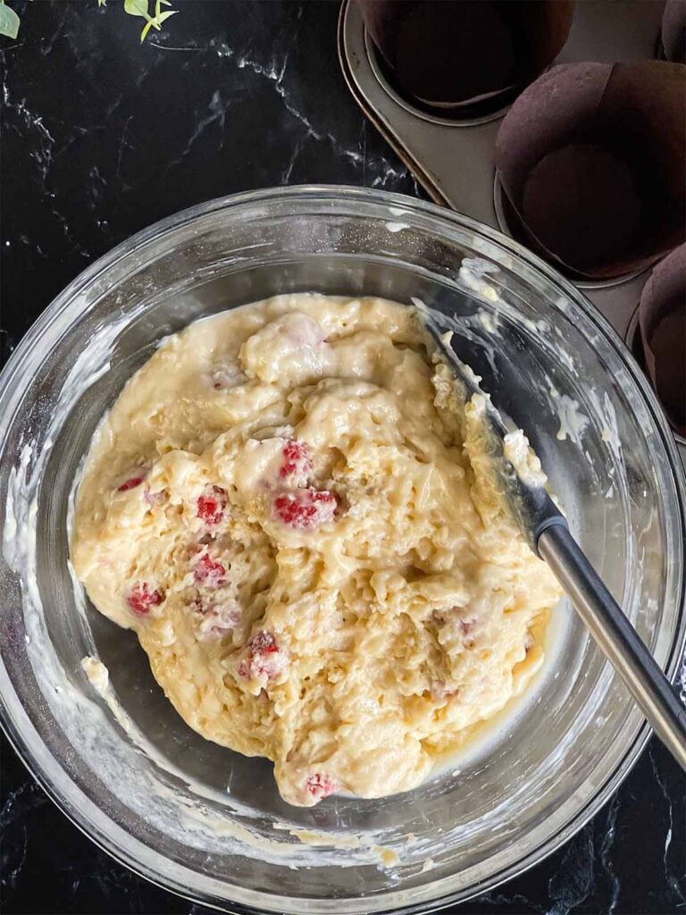 Raspberry muffin batter in a glass mixing bowl on a dark surface after resting for 15 minutes.
