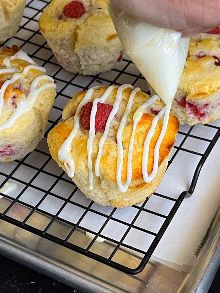 Lemon glaze being drizzled over the raspberry muffins on a wire rack over a parchment paper lined baking sheet.