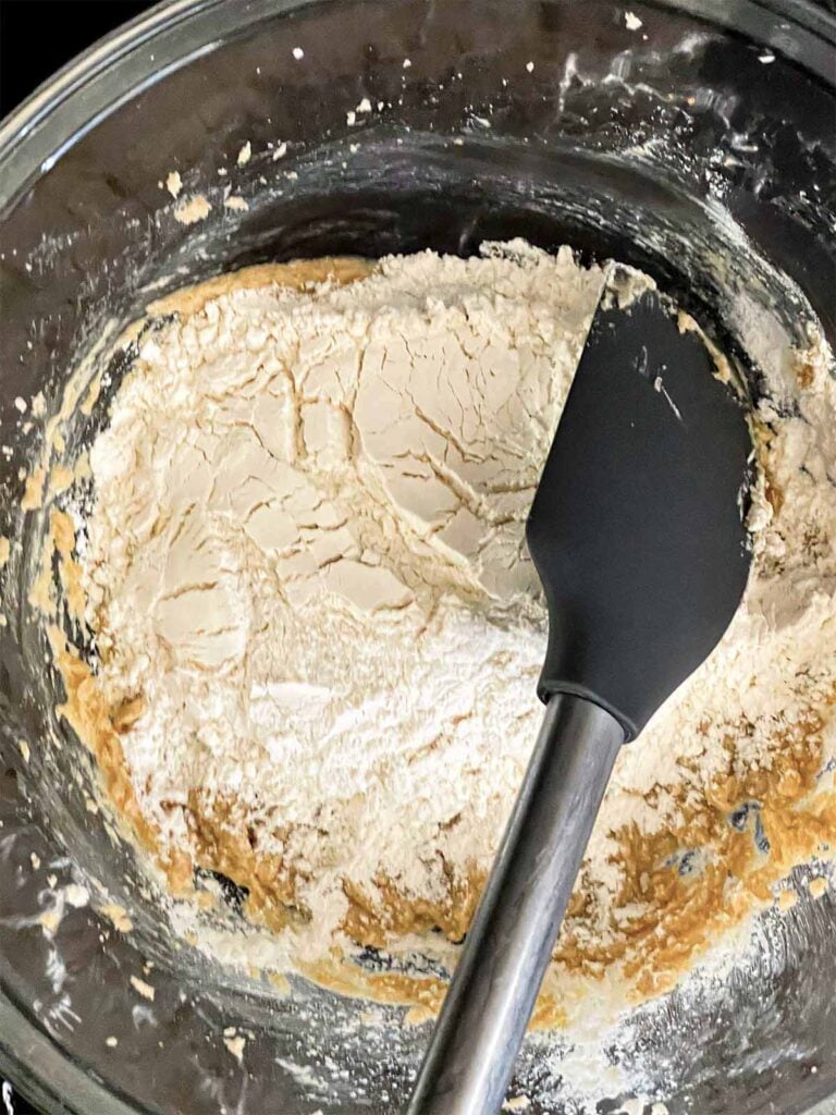 Dry ingredients add to the wet ingredients in a glass mixing bowl for almond joy cookies.