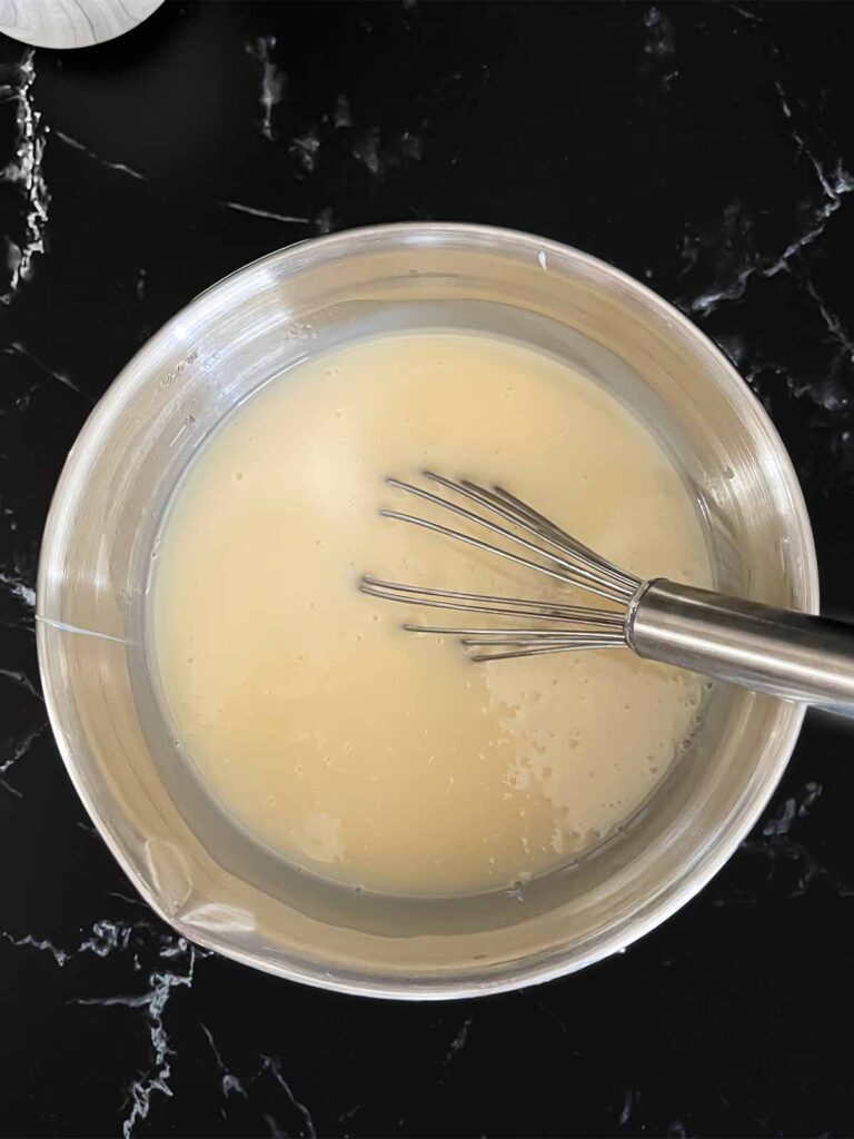 Cream of coconut whisked together with the sweetened condensed milk in a metal mixing bowl on a dark surface.