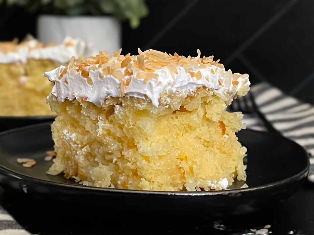 Coconut sheet cake slice on a dark plate on a dark surface.