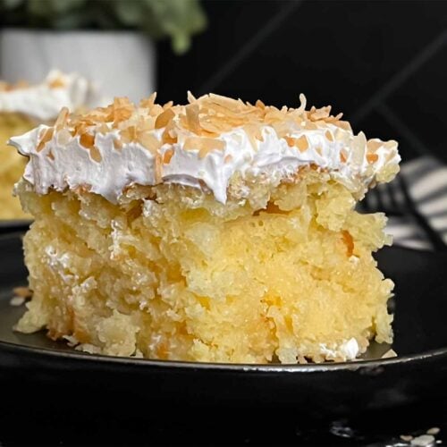 Coconut sheet cake slice on a dark plate on a dark surface.