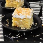 Coconut sheet cake slice on a dark plate on a dark surface.