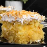 Coconut sheet cake slice on a dark plate on a dark surface.