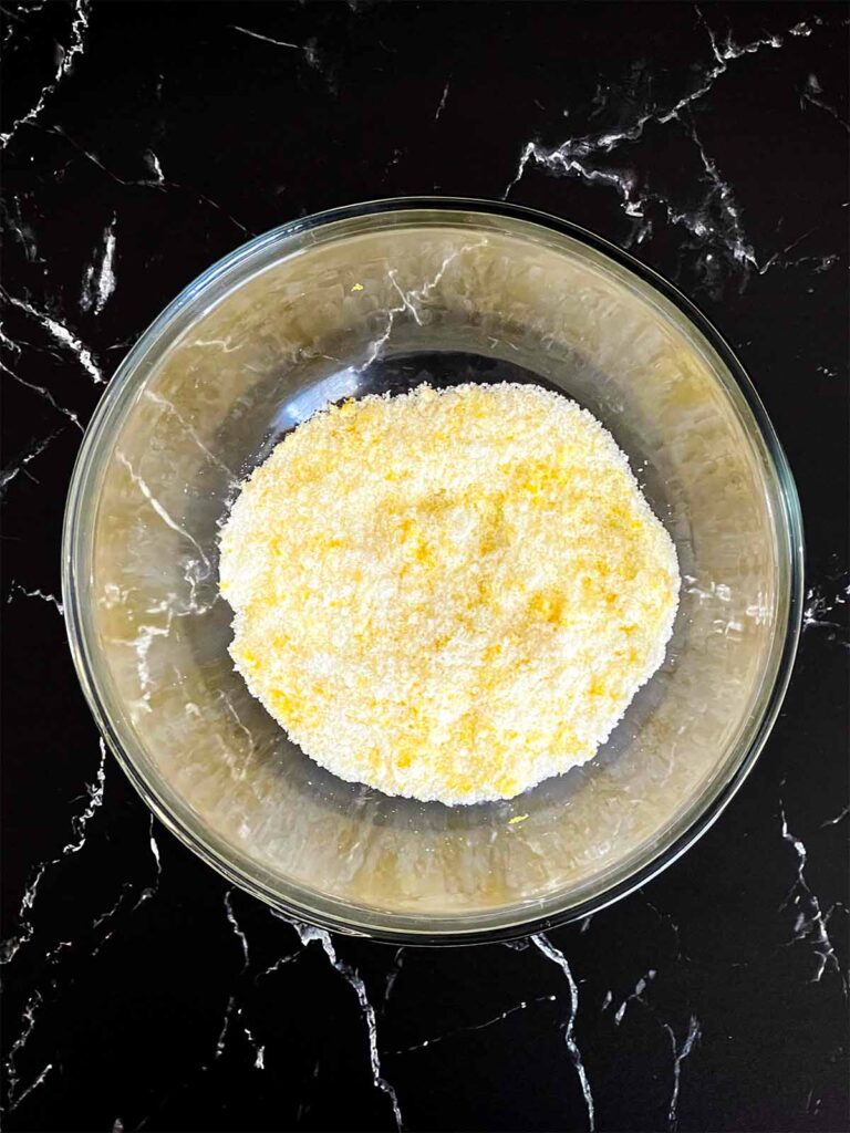 Lemon zest and granulated sugar rubbed together in a glass mixing bowl for lemon crumb cake recipe.