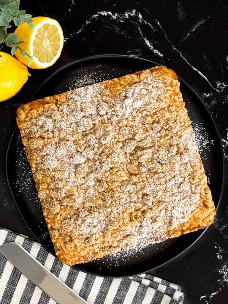 Baked lemon crumb cake dusted with powdered sugar on a dark plate on a dark surface.