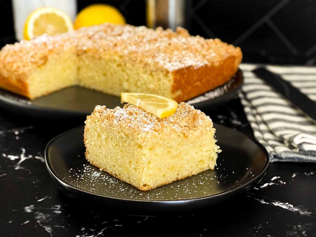 A slice of lemon crumb cake dusted with powdered sugar and garnished with a lemon slice on a dark plate on a dark surface.