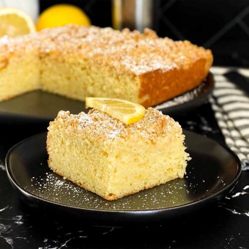 A slice of lemon crumb cake dusted with powdered sugar and garnished with a lemon slice on a dark plate on a dark surface.