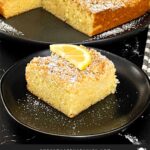 A slice of lemon crumb cake dusted with powdered sugar and garnished with a lemon slice on a dark plate on a dark surface.