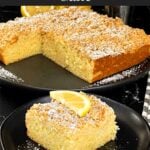 A slice of lemon crumb cake dusted with powdered sugar and garnished with a lemon slice on a dark plate on a dark surface.
