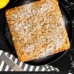 Lemon crumb cake dusted with powdered sugar on a dark plate on a dark surface.