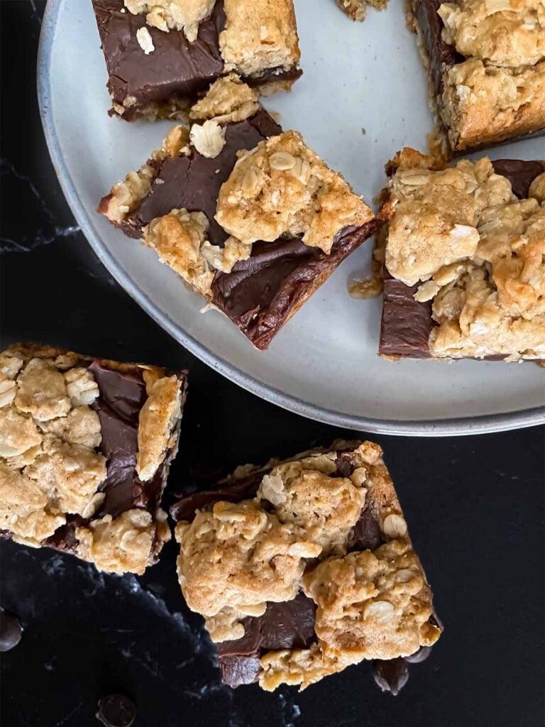 Oatmeal fudge bars on a dark surface in front of a plate of more bars.