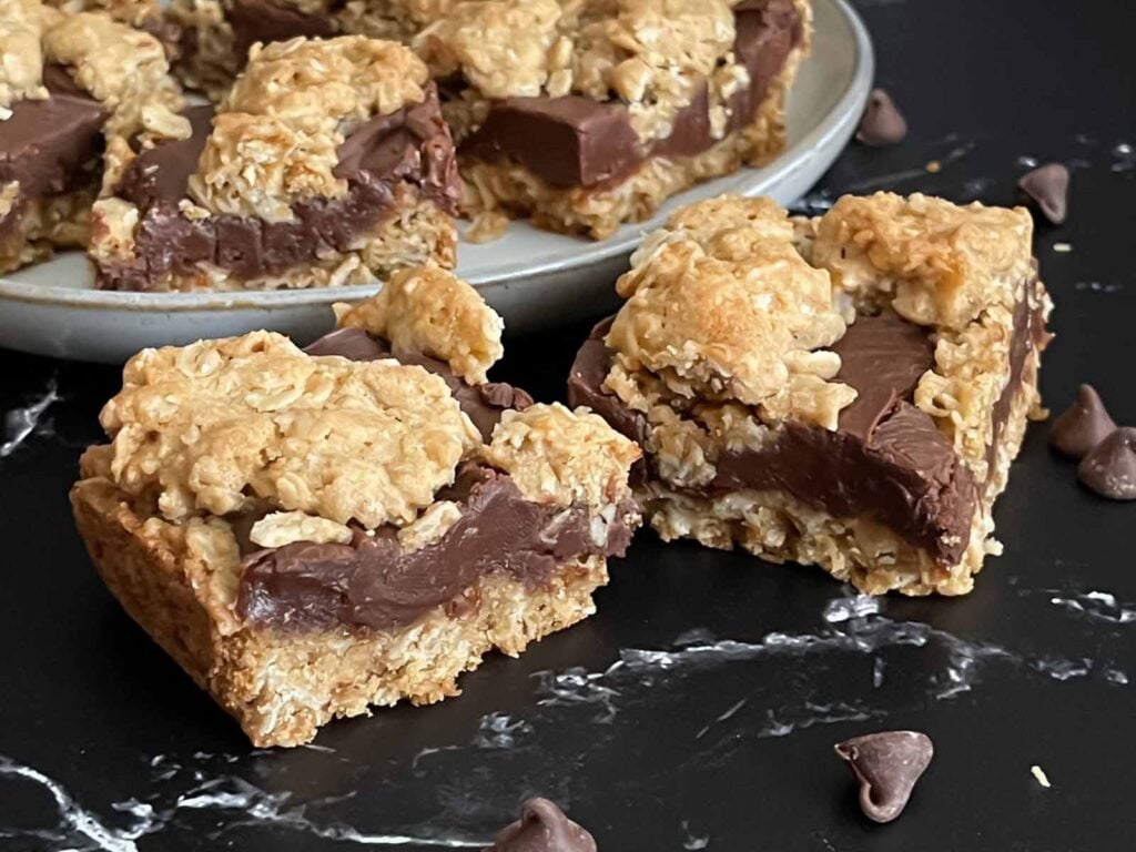 Oatmeal fudge bars on a dark surface in front of a plate of more bars.
