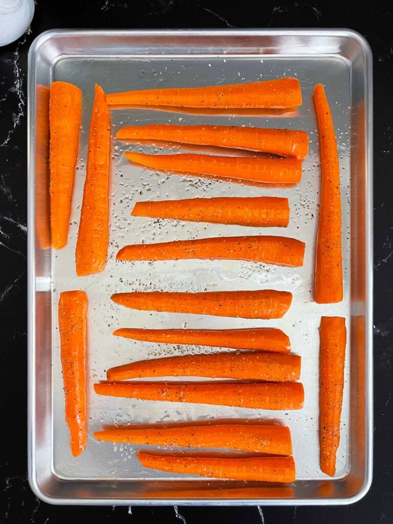 Raw whole carrots tossed in oil and seasonings on a metal rimmed baking sheet.
