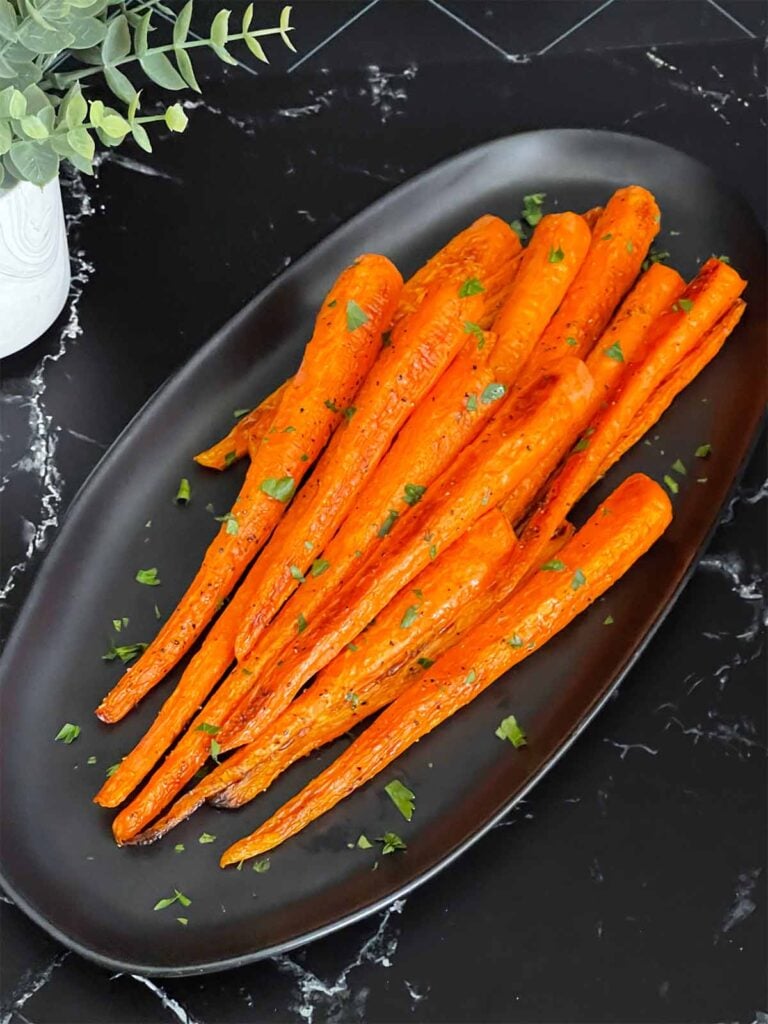 Roasted whole carrots on a dark plate garnished with chopped parsley.