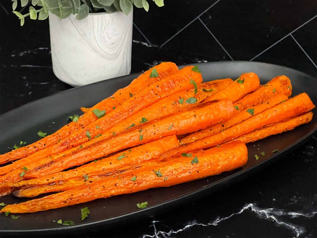 Roasted whole carrots on a dark plate garnished with chopped parsley.