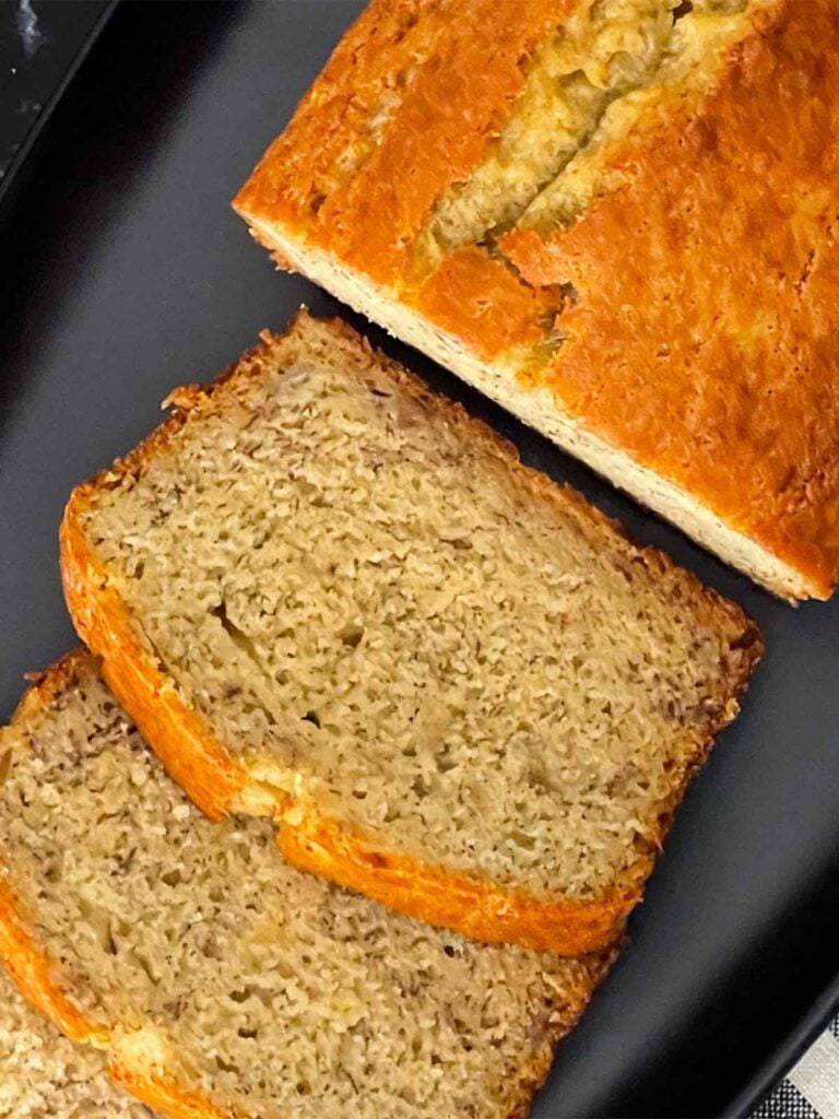 Slices of banana bread and the remaining loaf on a dark plate on a dark surface.