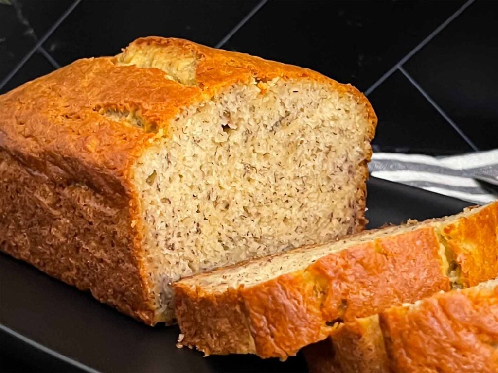 Slices of banana bread and the remaining loaf on a dark plate on a dark surface.