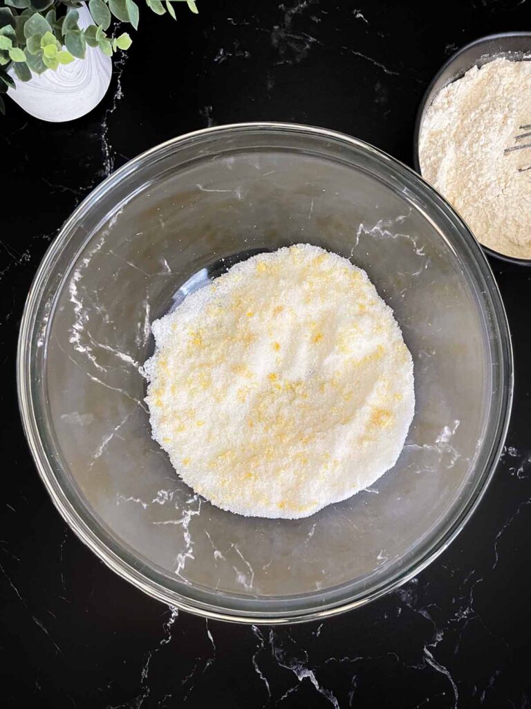 Granulated sugar and lemon zest rubbed together in a glass mixing bowl on a dark surface.