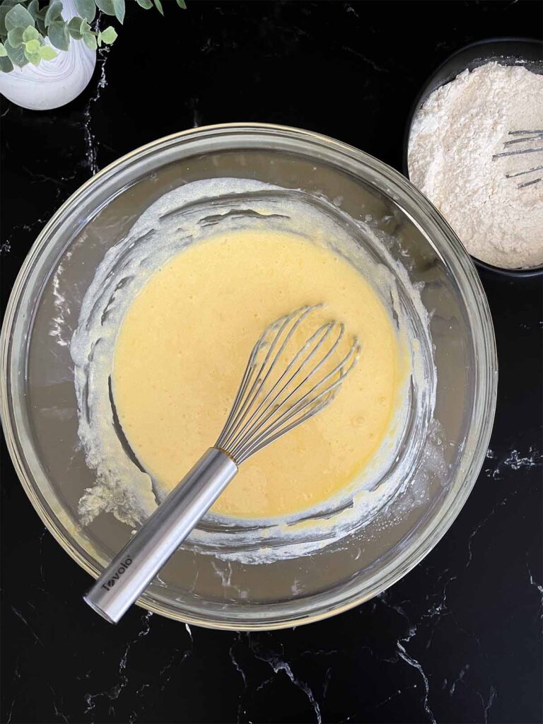 Wet ingredients for blueberry bread whisked together in a glass mixing bowl on a dark surface.