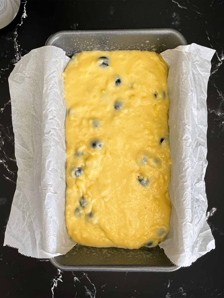 Blueberry bread batter in a parchment paper lined loaf pan on a dark surface.