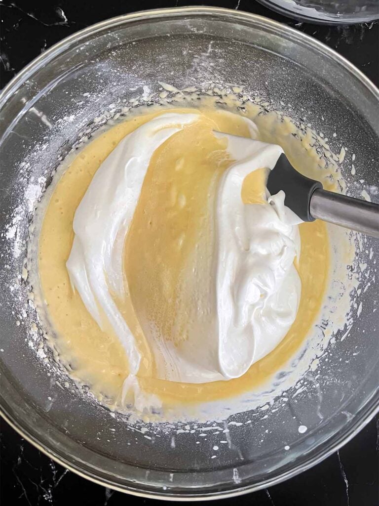 Whipped egg whites being gently folded into the coconut cake batter in a glass mixing bowl on a dark surface.
