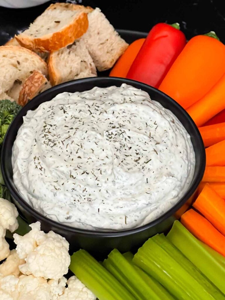 Dill dip in a small dark bowl surrounded by carrots, celery, cauliflower, broccoli, mini bell peppers and rye bread pieces on a dark plate.