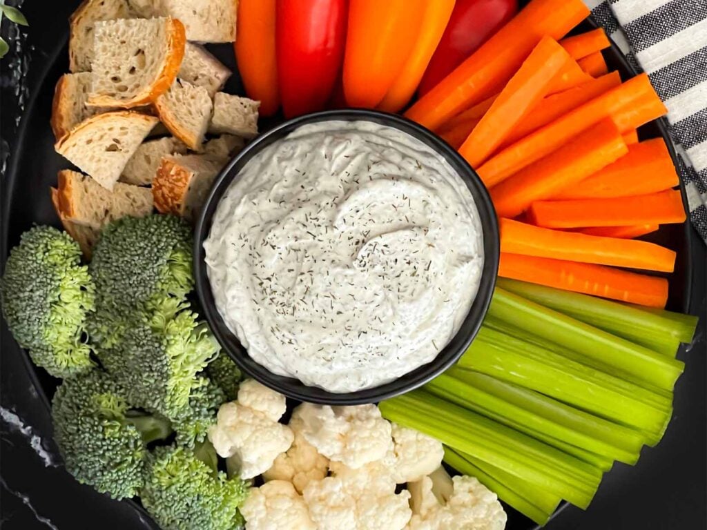 Dill dip in a small dark bowl surrounded by carrots, celery, cauliflower, broccoli, mini bell peppers and rye bread pieces on a dark plate.