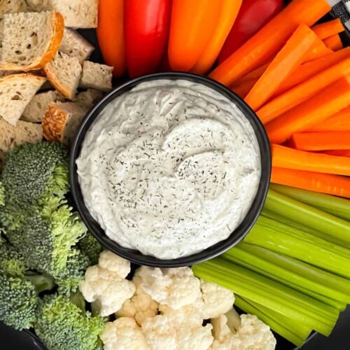 Dill dip in a small dark bowl surrounded by carrots, celery, cauliflower, broccoli, mini bell peppers and rye bread pieces on a dark plate.