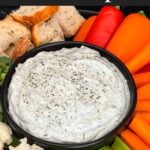 Dill dip in a small dark bowl surrounded by carrots, celery, cauliflower, broccoli, mini bell peppers and rye bread pieces on a dark plate.