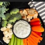 Dill dip in a small dark bowl surrounded by carrots, celery, cauliflower, broccoli, mini bell peppers and rye bread pieces on a dark plate.
