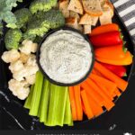 Dill dip in a small dark bowl surrounded by carrots, celery, cauliflower, broccoli, mini bell peppers and rye bread pieces on a dark plate.