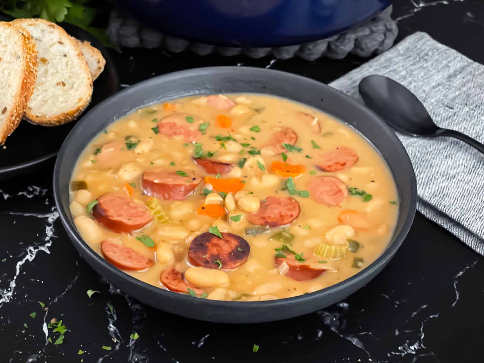 A bowl of creamy bean soup with sausage in a dark bowl on a dark surface.