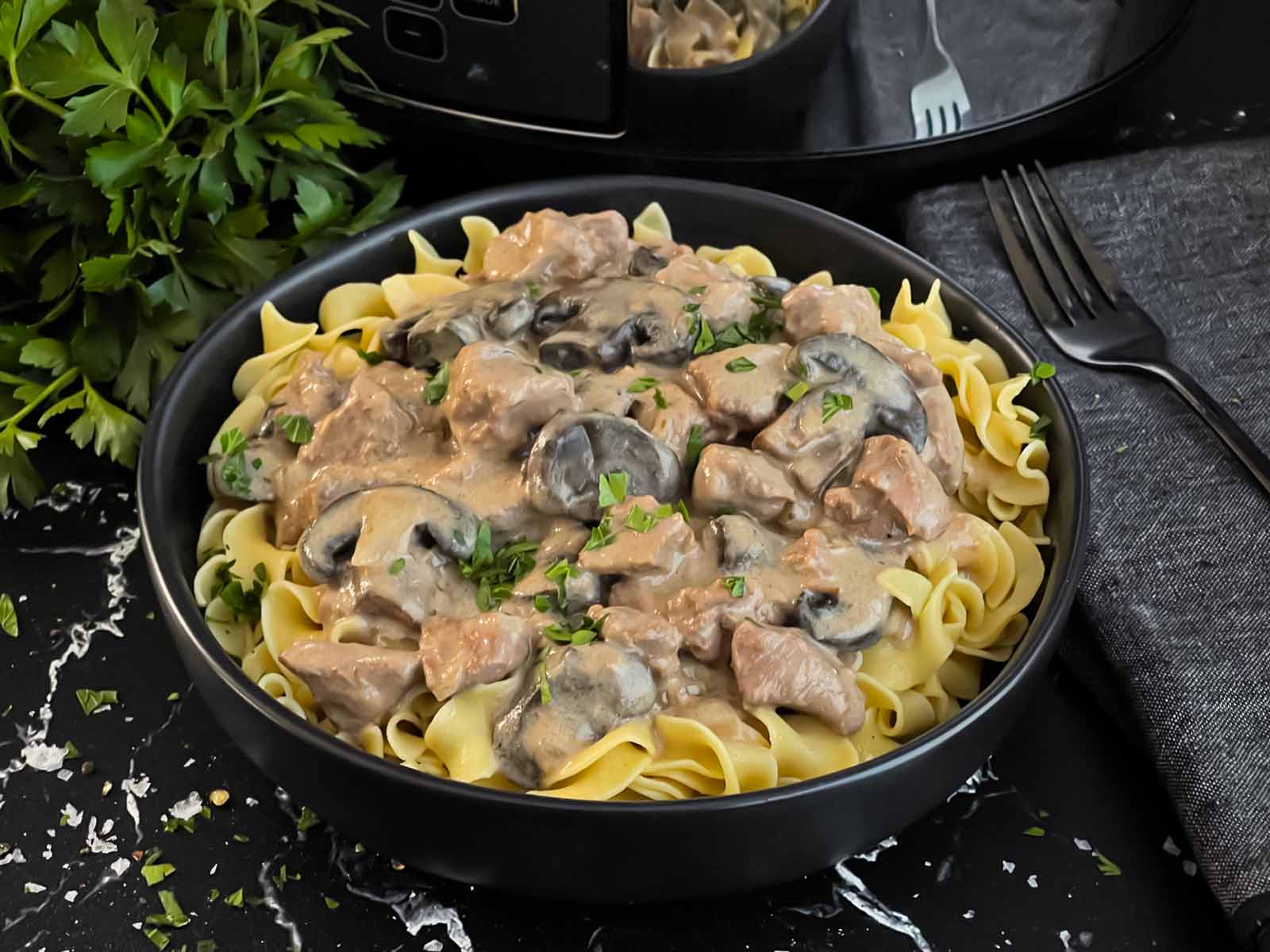 Slow cooker beef stroganoff over a bed of egg noodles in a dark bowl.
